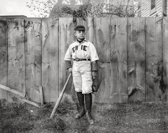 Photo showing: Erie Slugger -- Circa 1900-1910. Boy in 'Erie' baseball uniform standing by fence.